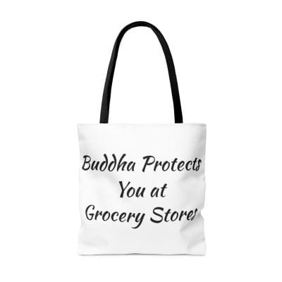 The Buddha Tote Bag (AOP) in white, featuring black handles, prominently showcases the phrase "Buddha Protects You at Grocery Stores" in bold black lettering.