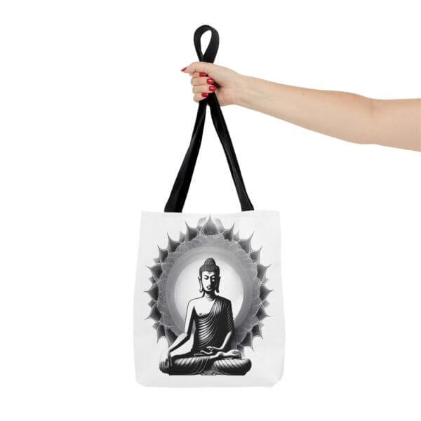 A hand holds the Buddha Tote Bag (AOP), which features a black-and-white design of a meditating figure against a circular pattern. A hand holds the Buddha Tote Bag (AOP), which features a black-and-white design of a meditating figure against a circular pattern.