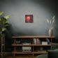 The "Blood Moon Over Ruins — Dystopian Cityscape Canvas, Stretched, 1.25"" hangs on a gray textured wall above a wooden shelf with books, an old radio, a vase, and a plant.